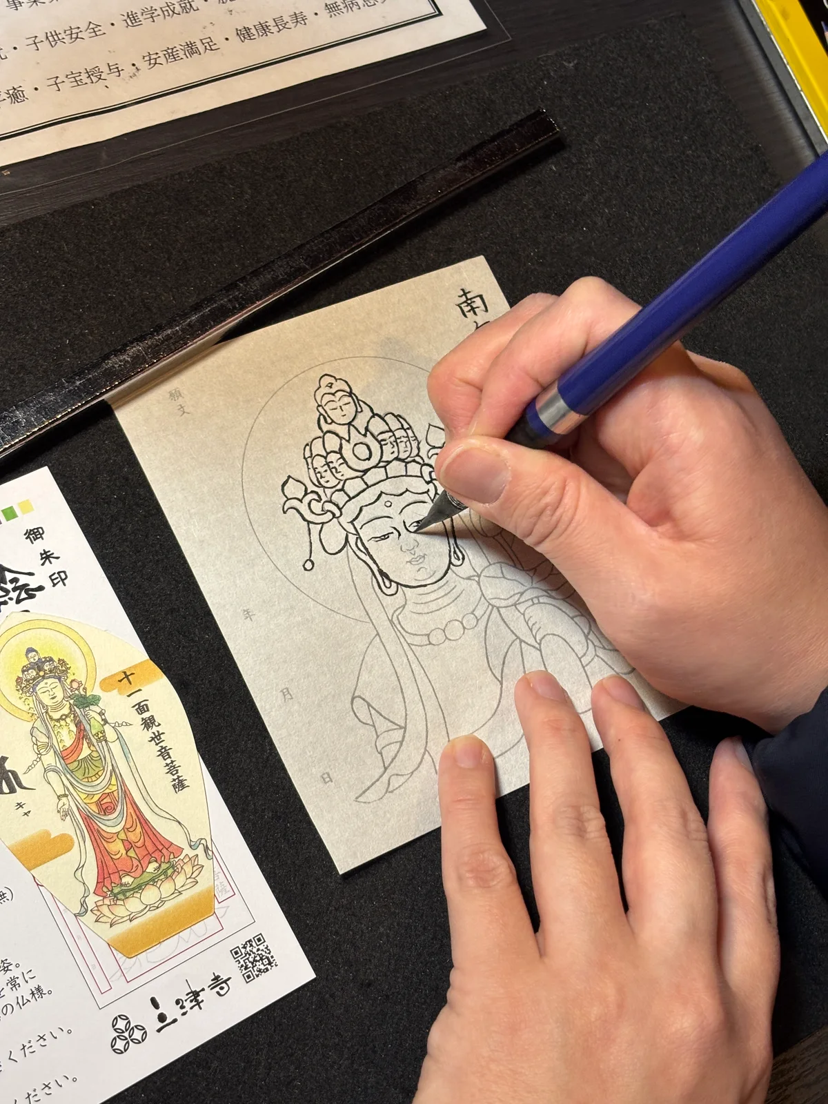 Hands using a brush pen to trace a Buddhist image on a Mitsutera eshakyo sheet